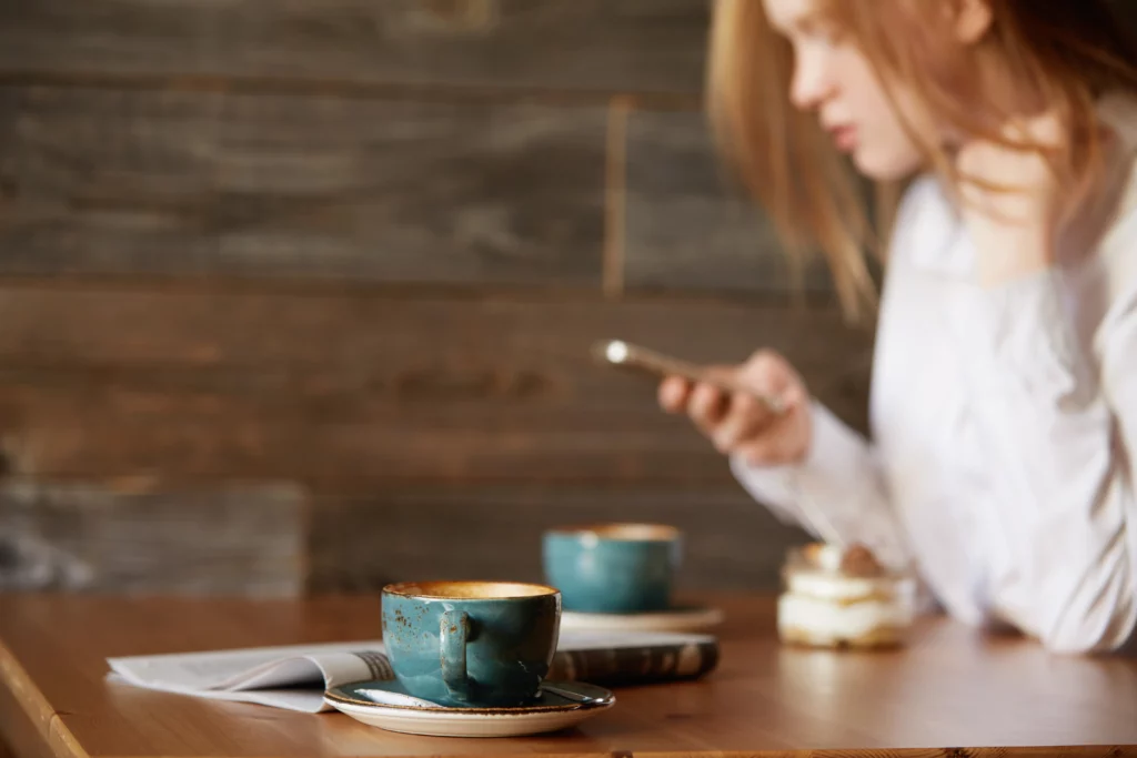 Tasse de thé en céramique bleue sur table en bois avec femme consultant son téléphone en arrière-plan pour page contact GreenSip